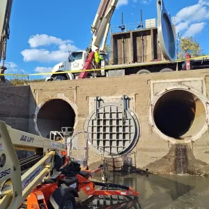 Truck-mounted crane lowering an 84-inch flap gate to the culvert headwall, one gate already hung