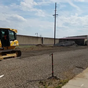 Tracked dozer on a fresh gravel working platform between the concrete floodwall and the railroad track, with rock and soil stockpiles ahead