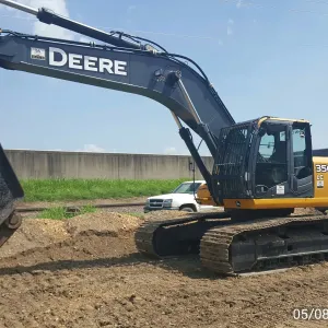 Larger tracked excavator with a hydraulic shear attachment on the dirt pad beside the railroad track and floodwall