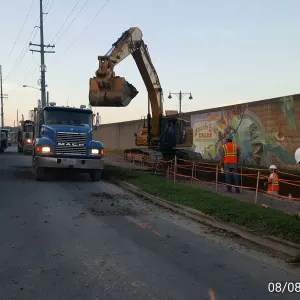 Excavator loading a dump truck in the street at dusk while a laborer rakes spoil beside the painted floodwall