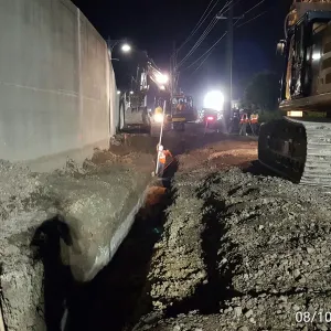 Narrow trench cut tight against the floodwall at night, with a worker down in the trench and excavators working the line