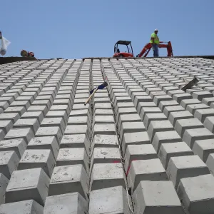Looking up a slope of placed articulated concrete block revetment toward a worker and compact excavator on the crest, with a cut-off saw set beside them