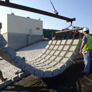 Crew steadying a suspended mat of articulated concrete block as it is lowered beside an already-placed section next to the outlet control building