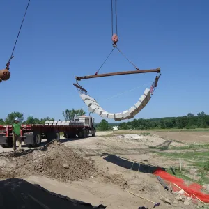 Crane lifting a slung mat of articulated concrete block off a flatbed truck, with the remainder of the load still on the trailer