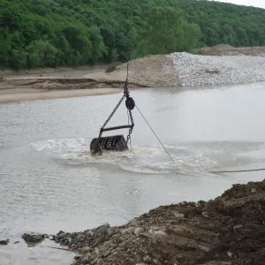 Clamshell bucket dropping into flowing muddy water at the edge of a rock fill, with an excavator working on the far bank