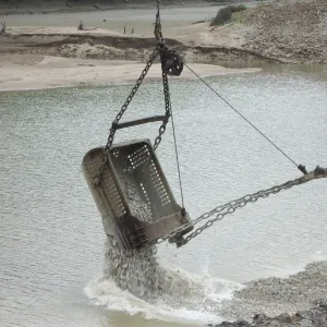 Clamshell bucket discharging crushed stone into shallow water beside a sand bar and stone bank