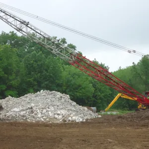 Lattice-boom crawler crane with a clamshell bucket working beside a large stockpile of shot rock
