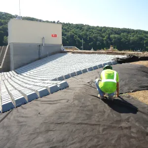 Worker kneeling with a tape measure on geotextile beside articulated concrete block fanned out from the outlet structure, concrete stairs and handrail at left