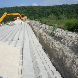 Long run of articulated concrete block down a slope beside an open excavated trench cut into rock, crew working at the far end