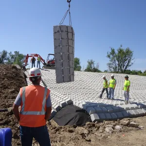 Crane holding a stacked bundle of blocks above the revetment while workers watch, geotextile turned up at the toe