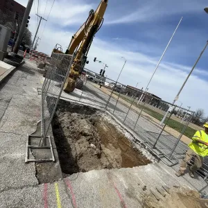 Open excavation at a curb line showing layered fill and native clay in the sidewall, with chain-link fence panels, orange barricades, and an excavator behind