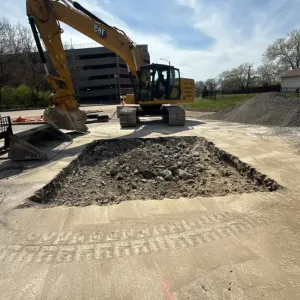 Excavator parked beside a broken-out pavement opening filled with rubble and spoil, with an aggregate stockpile at right