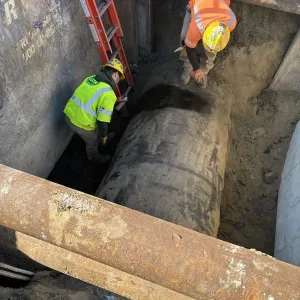 Two workers in a shored trench working alongside a large-diameter pipe, with steel struts crossing the trench and a ladder for access