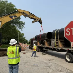 Excavator rigged to lift a section of large-diameter cement-lined pipe from a flatbed trailer on a tree-lined street, with two workers in reflective vests watching