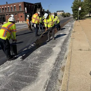 Crew brooming and applying tack coat along the joint between new asphalt paving and existing concrete pavement on a wide street