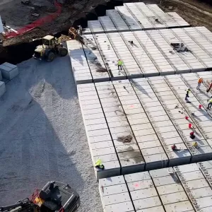 Aerial view of workers laying out and marking the tops of stenciled pre-placed crypt rows, with a compact tracked machine on the crypt deck, individual crypt units staged on a crushed-stone working pad at left, a wheel loader beside a white jobsite office trailer, and an excavator entering at the lower edge