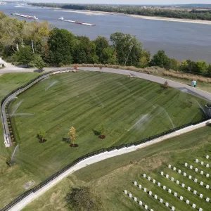 Aerial view of a completed sodded burial expansion panel with irrigation sprinklers running across the turf, bordered by a curving stone-faced retaining wall with black picket fence and a new asphalt loop road, with young landscape trees in mulch rings, existing headstone sections at left and lower right, and a barge tow moving on the Mississippi River beyond
