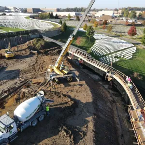 Aerial view of retaining wall construction with a rough-terrain crane lifting a suspended concrete bucket over the formed wall section, a concrete mixer truck and worker at lower left, an excavator and stacked steel beyond the spoil pile, and workers on the plank walkway beside the reinforcing cage, with existing headstone sections and hospital buildings in the background