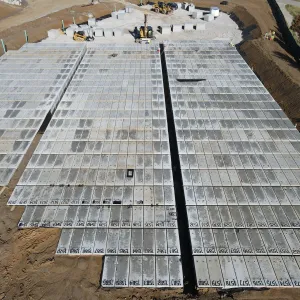Aerial view of two large blocks of numbered pre-placed crypts separated by a black-lined open drainage trench, with an excavator and skid steer working the staging pad at the top of frame, a surveyor with a tripod-mounted instrument at the right edge, and geotextile-covered excavation slopes on both sides