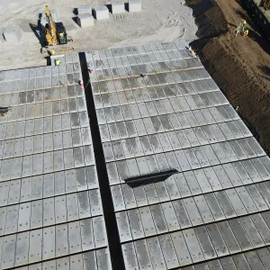 Close aerial view of the grid of pre-placed crypt lids with a black liner strip running between the two blocks, a worker's tool bag on the deck, an excavator handling crypts on the crushed-stone pad above, and a fabric-lined earth slope with silt fence along the right side