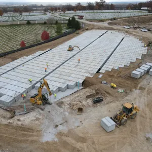 Aerial view of a nearly complete pre-placed crypt array with workers spaced across the deck, a compact excavator on the crypt tops, a larger excavator and a wheel loader carrying a crypt unit along the lower edge, staged crypts and lumber on the surrounding work pad, and existing headstone terraces beyond the fence line