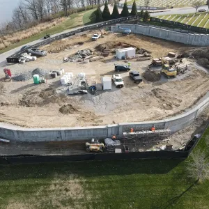 Aerial view of a walled work area in mid-construction with masons on rolling scaffold setting stone veneer along the outer face of the concrete retaining wall, and the enclosed pad filled with a concrete washout, portable toilet, aggregate stockpiles, a storage container, pickup trucks, a tracked excavator and stacked forming materials