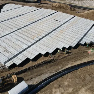 Aerial view of a very large completed block of pre-placed crypts with stepped end rows exposed, workers and a walk-behind compactor at the open edge, an excavator and skid steer working the backfill, a wheel loader hauling a crypt unit at the lower left, and a black-lined drainage swale crossing the foreground