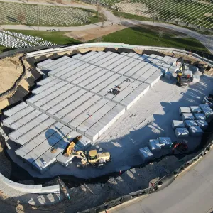 Aerial view of pre-placed crypts being set into a lined excavation, with a wheel loader positioning a unit at the lower left, a tracked excavator setting crypts at the upper right, dozens of individual crypts staged on the crushed-stone pad, silt fence along the perimeter, and existing headstone sections on the slope above