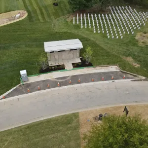 Aerial view of the completed restroom building with stone veneer walls and a standing-seam metal roof, a new concrete walk and freshly paved asphalt pull-off lined with traffic cones, a portable toilet and erosion control sock along the curb, and a newly set section of headstones on the lawn above