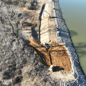Aerial view of a long-reach excavator, a dozer and pickup trucks working a stripped embankment crest with riprap along the waterline
