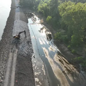 Excavator spreading light-colored bedding stone along the riverside slope in low morning sun