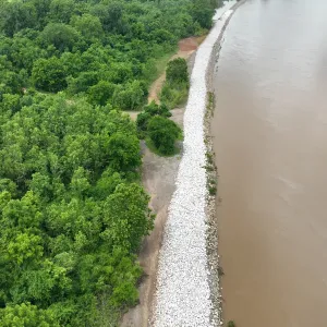 Aerial view of a continuous band of newly placed riprap along the riverbank with dense woods on the landside